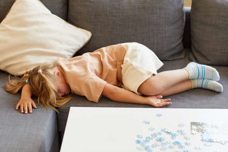 Full length portrait of cute girl with down syndrome falling asleep on sofa while playing board...