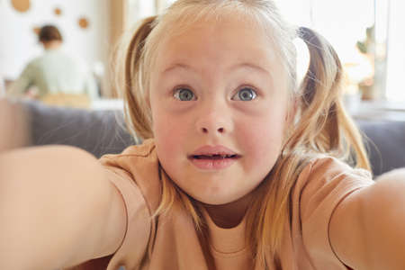 Close up portrait of cute little girl with down syndrome taking selfie and holding camera at home, copy spaceの写真素材