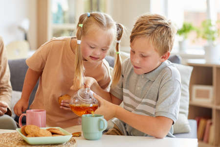 Portrait of cute girl with down syndrome enjoying tea with family while sitting in sunlit living room at home, copy spaceの写真素材