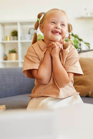 Vertical portrait of cute girl with down syndrome laughing happily while sitting on couch in sunlit roomの写真素材