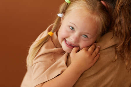 Close up of mother holding happy daughter with down syndrome while standing against plain brown...