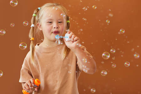 Waist up portrait of cute girl with down syndrome blowing bubbles while posing against brown...