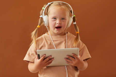 Waist up portrait of cute girl with down syndrome holding digital tablet and looking at camera while...