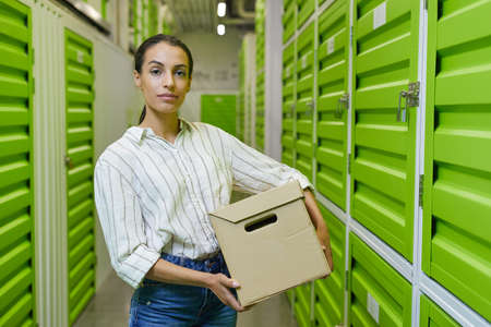 Waist up portrait of young woman holding cardboard box standing in self storage facility and looking at camera, copy spaceの写真素材