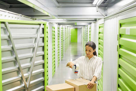 Wide angle portrait of young woman packing boxes with tape gun while standing by self storage unit,...