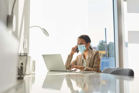 Portrait of mixed-race businesswoman wearing mask and speaking by smartphone while working in white...