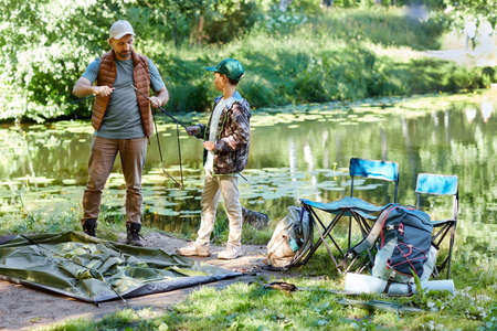 Full length portrait of father and son setting up tent together while camping by lake in nature,...