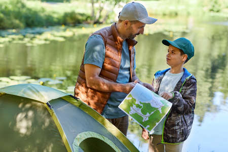 Waist up portrait of father and son looking at map while enjoying camping together by lake, copy space