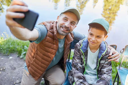 High angle portrait of loving father and son taking selfie photo via smartphone while enjoying camping trip togetherの写真素材