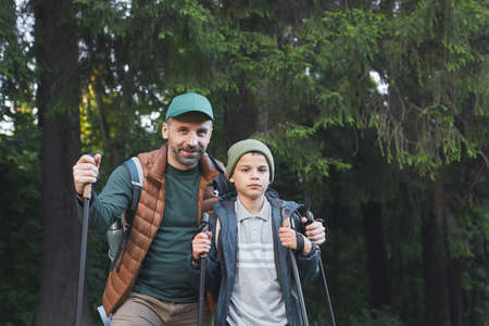 Waist up portrait of happy father and son hiking together and looking at camera while walking in...