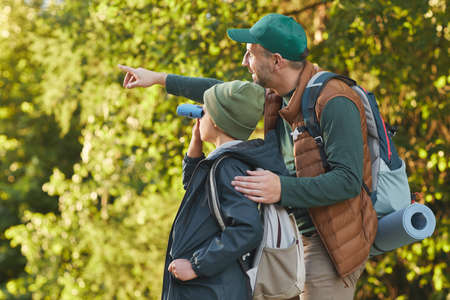 Portrait of happy father and son hiking together and pointing away while walking in forest with...