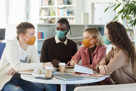 Multi-ethnic group of young people wearing masks while studying together at table in college library, copy spaceの写真素材