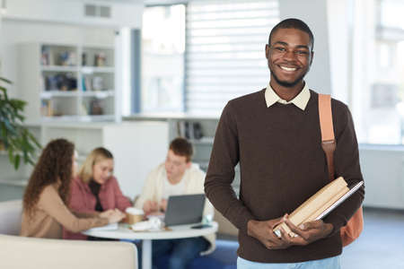 Waist up portrait of smiling African teenage boy looking at camera and smiling while standing in college library with people working in background, copy spaceの写真素材