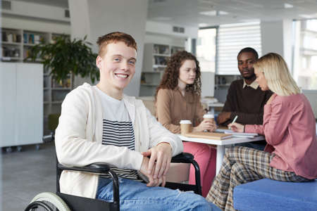 Portrait of smiling red haired boy using wheelchair studying with group of students in college...