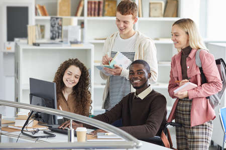 Multi-ethnic group of students using telecommunication equipment while studying in college library,...