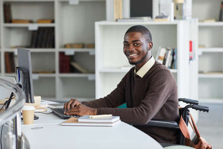 Portrait of disabled African man using computer and smiling at camera while studying in college library, copy spaceの写真素材