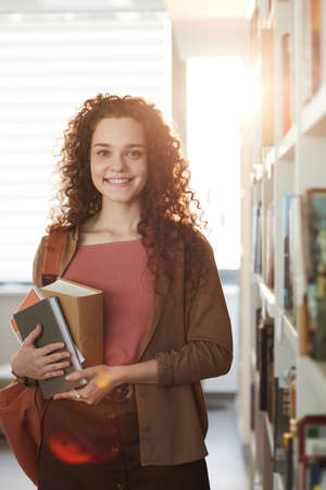 Vertical waist up portrait of young woman holding books and smiling at camera while standing in school libraryの写真素材