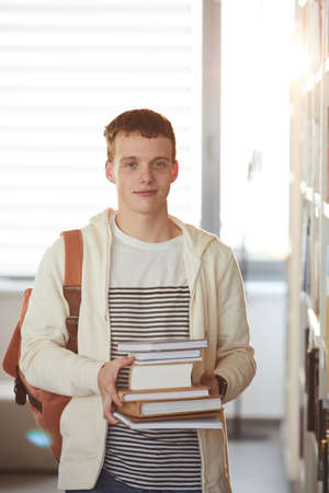 Vertical waist up portrait of young man holding books and smiling at camera while standing in school libraryの写真素材