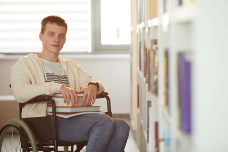 Warm toned portrait of young man using wheelchair in school while looking at camera in library lit...