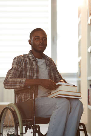 Vertical warm toned portrait of young African-American man using wheelchair in school while looking...