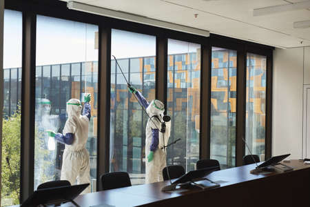 Wide angle portrait of two workers wearing hazmat suits disinfecting office windows in conference room, copy spaceの写真素材