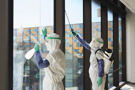 Portrait of two workers wearing hazmat suits disinfecting office windows in sunlight, copy space