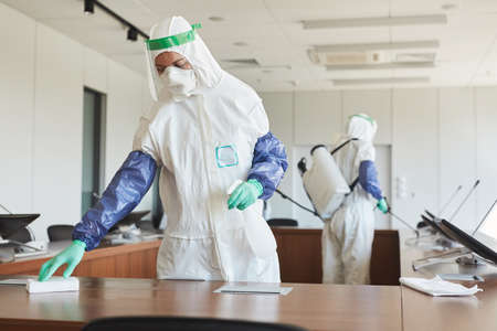 Portrait of two sanitation workers wearing hazmat suits cleaning and disinfecting conference room in office, copy spaceの写真素材