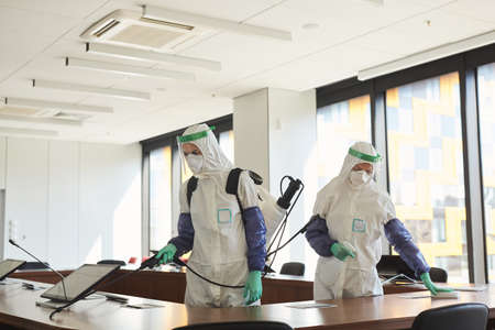 Wide angle portrait of two sanitation workers wearing hazmat suits cleaning and disinfecting...
