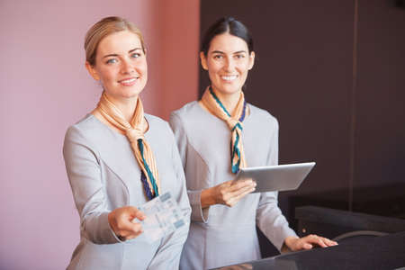 Portrait of two smiling flight attendants handing tickets to passenger while standing at check in desk in airport, copy spaceの写真素材