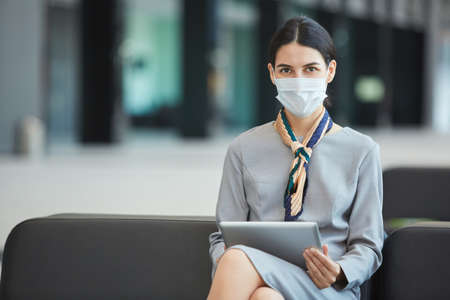 Portrait of female stewardess wearing mask and looking at camera while sitting on couch in waiting lounge of airport, copy spaceの写真素材