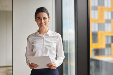 Waist up portrait of smiling businesswoman looking at camera while standing by window in office,...