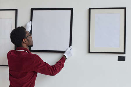 Back view at African-American man hanging frames on wall while planning art gallery or exhibition, copy spaceの写真素材
