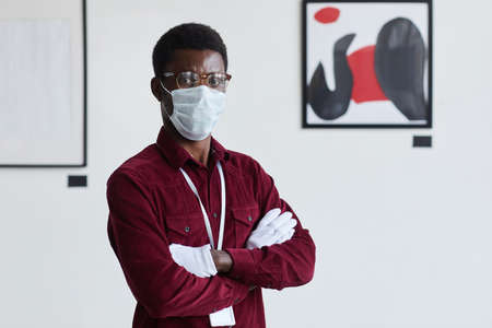 Waist up portrait of African-American man wearing mask looking at camera while standing with arms crossed against modern graphic paintings in art gallery, copy spaceの写真素材