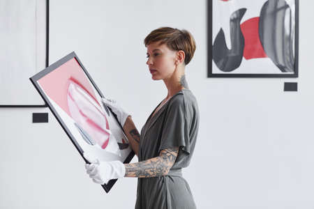 Waist up portrait of tattooed creative woman holding painting while planning art gallery exhibition, copy space