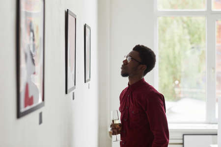 Side view portrait of young African-American man looking at paintings and holding champagne glass at art gallery opening, copy spaceの写真素材