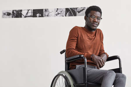 Minimal portrait of young African-American man using wheelchair and looking at camera while posing against white wall in modern art gallery, copy spaceの写真素材