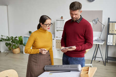 Waist up portrait of two architects using digital tablet while working on blueprints together in...