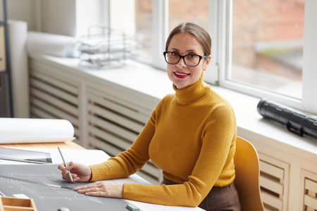 Portrait of smiling female architect drawing blueprints and plans while working at desk in office...