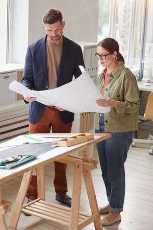 Vertical full length portrait of two architects holding blueprints and discussing work while standing by drawing desk in officeの写真素材