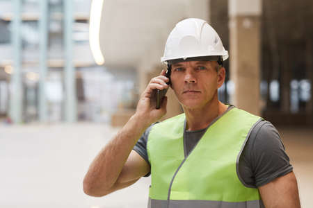 Portrait of professional construction worker speaking by phone and looking at camera while standing...