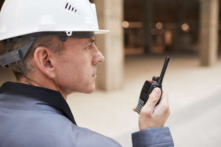 Side view portrait of mature worker speaking by walkie-talkie while supervising work at construction site or in industrial workshop, copy spaceの写真素材