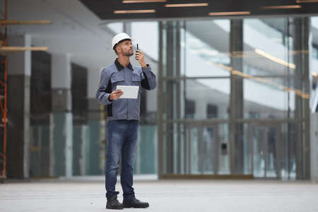 Full length portrait of mature worker speaking by walkie-talkie while supervising work at...