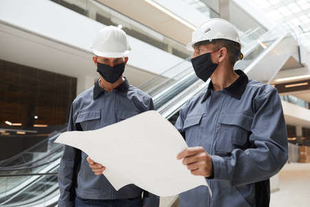 Waist up portrait of two construction workers wearing masks and discussing plans while standing in shopping mall or office buildingの写真素材