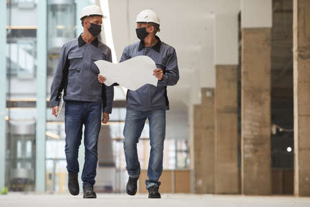 Full length portrait of two workers wearing masks and holding plans while walking towards camera at construction site, copy spaceの写真素材