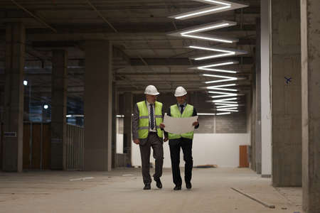 Full length portrait of two business people wearing hardhats and holding plans while walking towards camera at construction site, copy spaceの写真素材