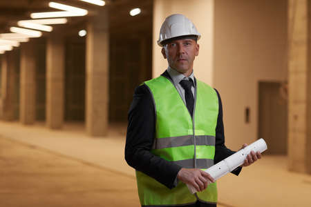Waist up portrait of mature businessman wearing hardhat and looking at camera while standing at...