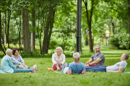 Wide shot of modern senior people spending summer morning together relaxing on grass in park after exercisingの写真素材