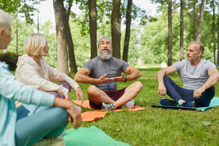 Stylish bearded yoga trainer sitting on mat demonstrating his senior clients how to do breath...