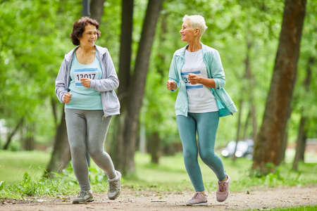 Full body shot of two active senior women running marathon race in city park talking to each other