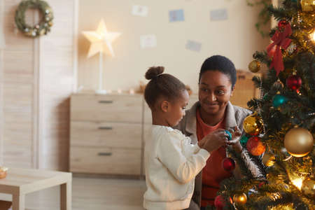 Side view portrait of cute African-American girl decorating Christmas tree with smiling happy mom in cozy home interior, copy spaceの写真素材